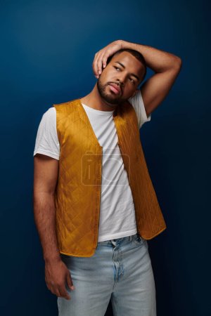 young handsome man in yellow vest posing on dark blue backdrop putting arm on his head, fashion