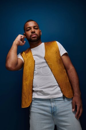 handsome stylish man in yellow vest posing with raised fist on dark blue backdrop, fashion concept