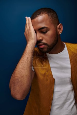 handsome african american man posing on dark blue backdrop with his palm on face, fashion concept