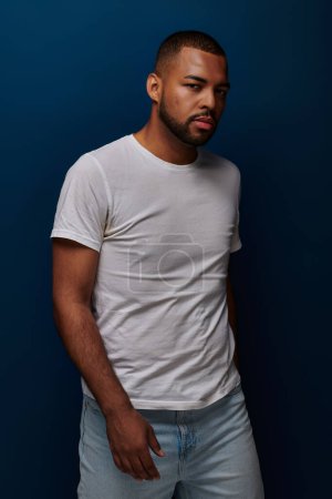 handsome young man in white t-shirt and jeans posing on blue backdrop looking away, fashion concept