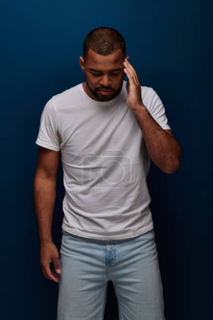 young man in stylish attire posing on dark blue backdrop with hand near forehead, fashion concept