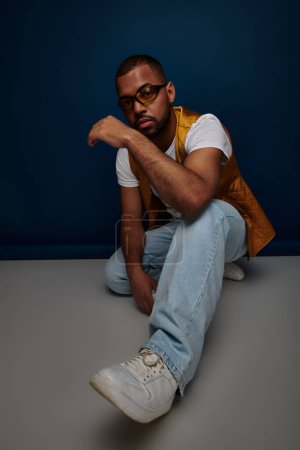 Photo for Handsome young man in vest and jeans squatting and posing on dark blue backdrop, fashion concept - Royalty Free Image