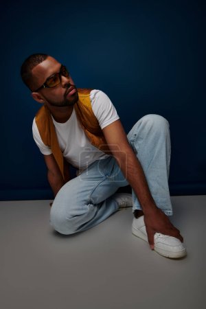 handsome stylish man in yellow vest and sunglasses sitting on floor looking away, fashion concept