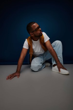trendy man in casual stylish outfit posing on floor looking away, hand to foot, fashion concept