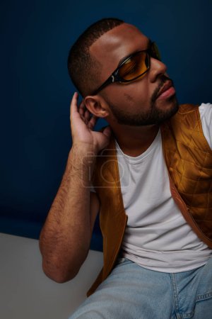 Foto de Primer plano afroamericano hombre en gafas de sol en azul oscuro telón de fondo mirando hacia otro lado, concepto de moda - Imagen libre de derechos