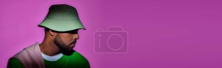 Photo for Young man in panama with earring posing in profile on purple backdrop, fashion concept, banner - Royalty Free Image