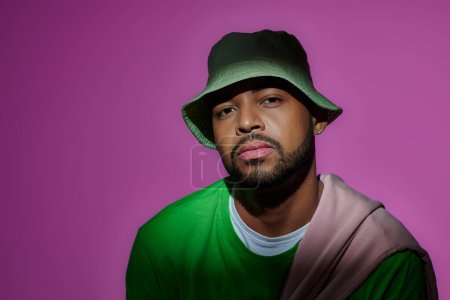 good looking man in green panama with earring on purple backdrop looking at camera, fashion concept