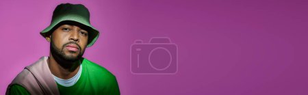 handsome bearded man in green panama on purple backdrop looking at camera, fashion concept, banner