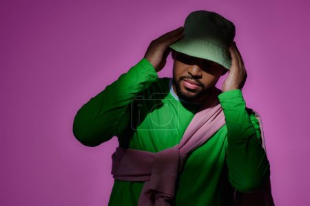 gut aussehender bärtiger Mann in grünem Sweatshirt, der sein grünes Panama vor violettem Hintergrund berührt, Mode