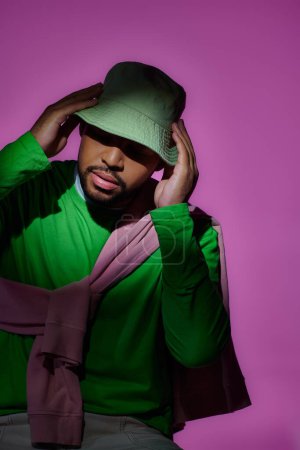 stylischer afrikanisch-amerikanischer Mann in grünem Sweatshirt, der sein Panama vor violettem Hintergrund berührt, Mode