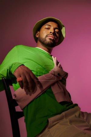joven guapo en panama verde sentado en la silla y mirando a la cámara, concepto de moda