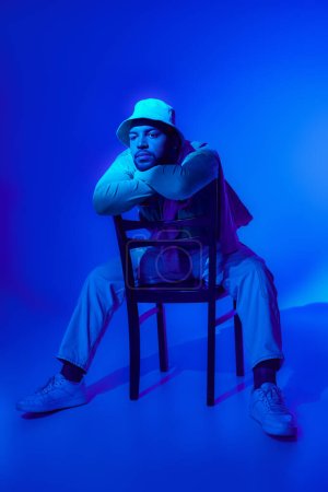 stylish african american in casual outfit sitting on chair surrounded by blue light, fashion concept