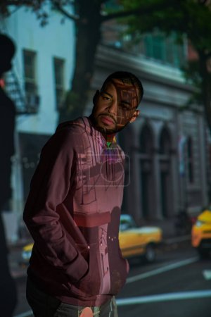 Foto de Bien parecido afroamericano en sudadera con capucha rosa posando en proyector digital luces, concepto de moda - Imagen libre de derechos