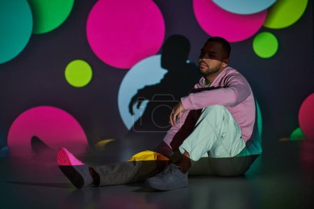 Foto de Modelo masculino atractivo con barba sentado en el suelo en luces proyectores digitales, concepto de moda - Imagen libre de derechos