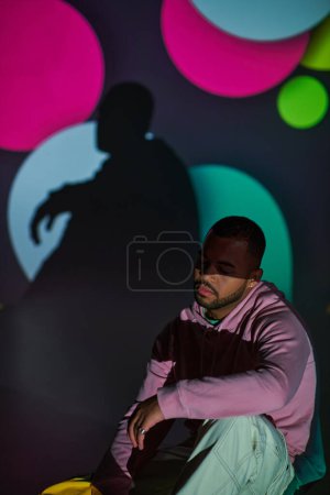 Téléchargez les photos : Bel homme en sweat à capuche rose assis sur le sol dans des projecteurs numériques, concept de mode - en image libre de droit
