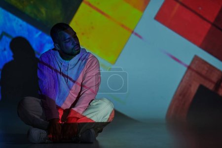 trendy man sitting on floor with crossed legs in pink hoodie in projector lights, fashion concept