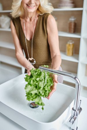 abgeschnittene Ansicht einer lebenslustigen Frau mittleren Alters mit blonden Haaren, die frischen Salat in der Spüle wäscht