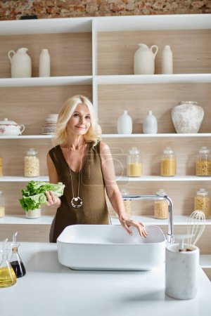 Foto de Atractiva mujer vegetariana de mediana edad con cabello rubio lavando lechuga fresca en fregadero de cocina - Imagen libre de derechos