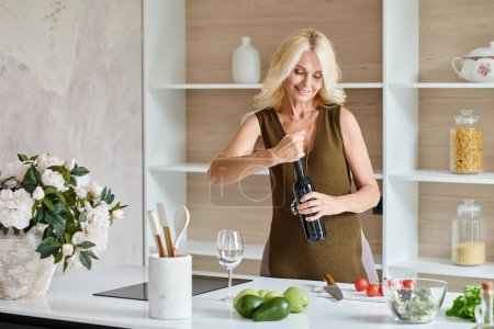 joyful middle aged woman with blonde hair opening bottle of wine near fresh vegetables on countertop