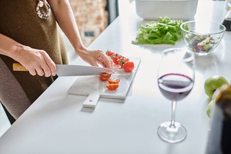 tiro parcial de mujer cortando tomates cherry cerca de un vaso de vino tinto y lechuga en un tazón transparente
