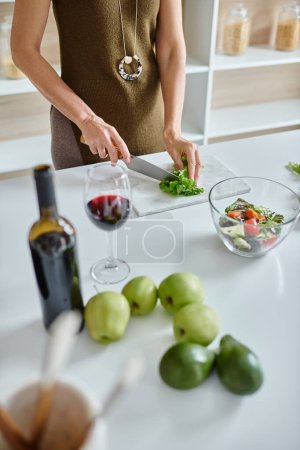 Foto de Mujer recortada cortando lechuga fresca y haciendo ensalada de verduras cerca de un vaso de vino tinto en la encimera - Imagen libre de derechos