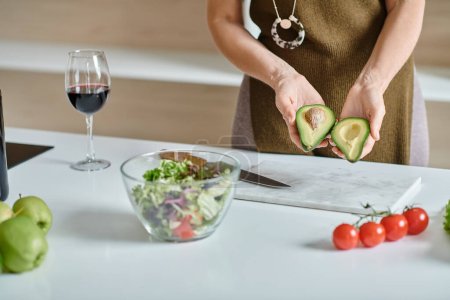 cropped woman holding fresh avocado halves near salad in bowl and glass of red wine on countertop