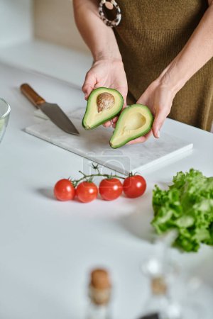 beschnittene Frau hält frische Avocadohälften in der Nähe von Kirschtomaten, Salat und Messer auf Arbeitsplatte
