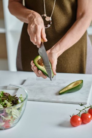 abgeschnittene Frau schneidet frische Avocado halb in der Nähe von Kirschtomaten und Salat in Schüssel, Hausmannskost