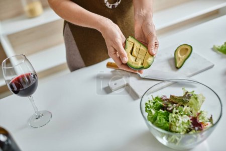 Nahaufnahme einer beschnittenen Frau mit geschnittener Avocado in der Nähe von Salat in Schüssel und Glas Rotwein