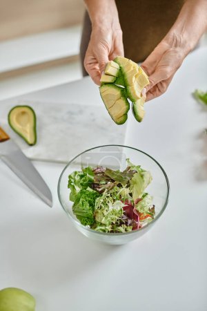 close up of cropped woman holding sliced ripe avocado near lettuce in bowl and glass of red wine