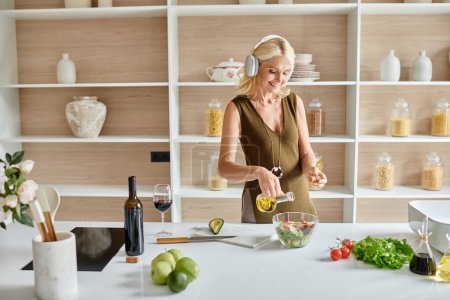 cheerful middle aged woman in wireless headphones making salad near bottle and glass of red wine