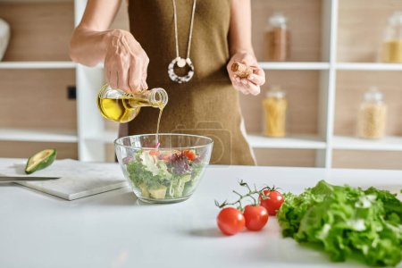 abgeschnittene Aufnahme einer Frau, die Olivenöl in eine Glasschüssel mit Salat in der Nähe von Gemüse auf der Arbeitsplatte gießt