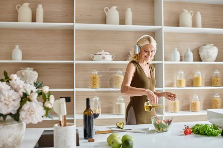 glückliche Frau mittleren Alters in drahtlosen Kopfhörern, die Salat in der Nähe von Flasche und Glas Rotwein zubereitet