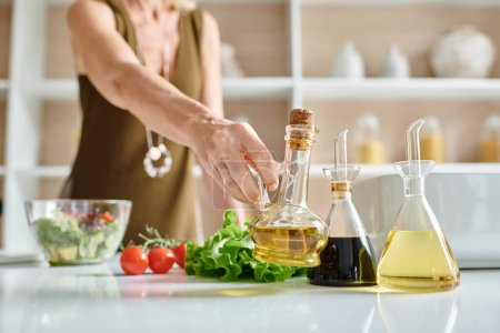 abgeschnittene Ansicht einer Frau, die eine Glasflasche mit Olivenöl nimmt, während sie in der Küche Salat zubereitet