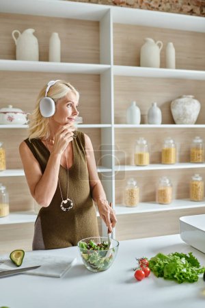 Unbekümmerte Frau mittleren Alters in drahtlosen Kopfhörern hört Musik und mischt Salat in der Küche