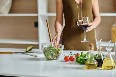 femme cultivée tenant un verre de vin rouge et debout près de la salade fraîchement préparée dans un bol transparent