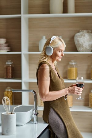 mujer de mediana edad en auriculares inalámbricos con vaso de vino tinto en la cocina moderna