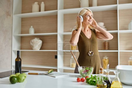 glückliche Frau mittleren Alters mit drahtlosen Kopfhörern, die neben frischen Zutaten und Schüssel in der Küche steht