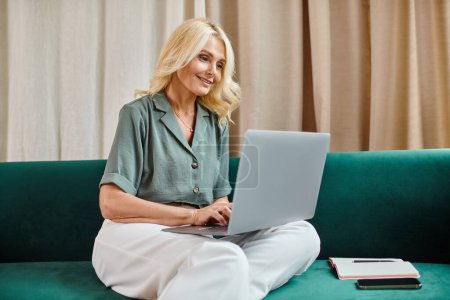 alegre mujer de mediana edad con el pelo rubio usando el ordenador portátil mientras está sentado en el sofá, trabajo remoto