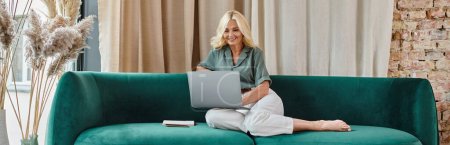 mujer de mediana edad relajada con el pelo rubio usando el ordenador portátil mientras está sentado en el sofá, el trabajo desde el banner del hogar