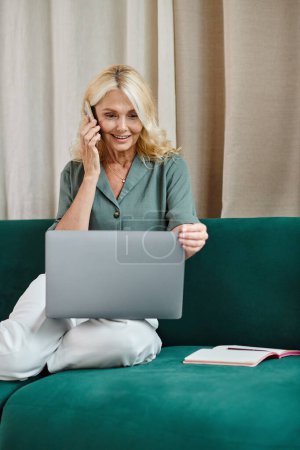Téléchargez les photos : Femme d'âge moyen avec des cheveux blonds parlant sur smartphone et utilisant un ordinateur portable, assis sur le canapé - en image libre de droit