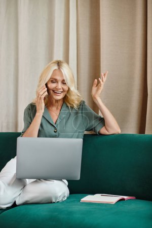 fröhliche Frau mittleren Alters mit blonden Haaren, die am Smartphone spricht und Laptop auf dem Sofa benutzt