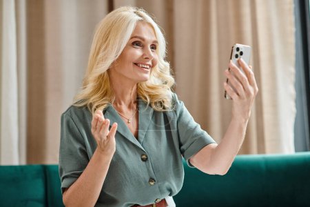glückliche Frau mittleren Alters mit blonden Haaren hat Videoanruf auf dem Smartphone und lächelt im Wohnzimmer