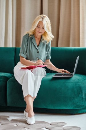 organized middle aged woman using laptop and taking notes while sitting on sofa in living room