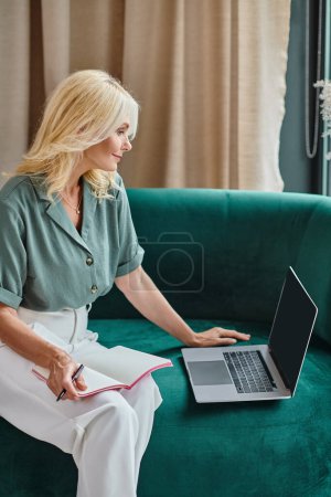 Téléchargez les photos : Attrayant femme d'âge moyen en utilisant un ordinateur portable et assis sur le canapé avec ordinateur portable sur ses tours - en image libre de droit