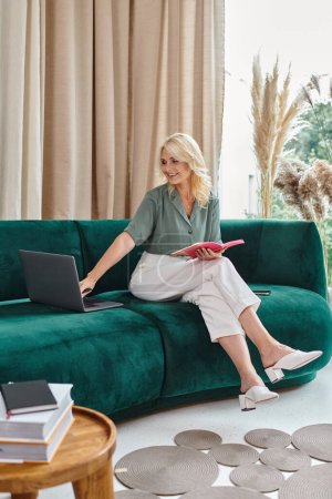 pleased middle aged woman using laptop and holding notebook while sitting on sofa in living room