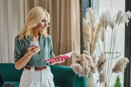 blonde middle aged woman looking at notes in notebook and holding smartphone while standing at home