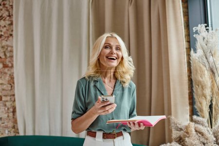aufgeregte Frau mittleren Alters mit blonden Haaren nutzt Smartphone und hält Notizbuch im Wohnzimmer