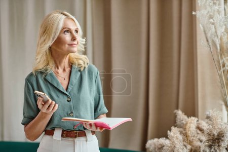 schöne Frau mittleren Alters mit blonden Haaren, Smartphone in der Hand und Notizbuch im Wohnzimmer
