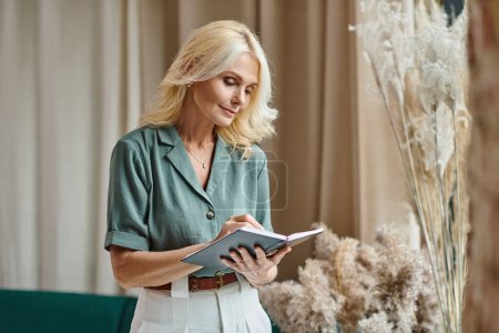 Photo for Beautiful middle aged businesswoman with blonde hair taking notes while holding notebook at home - Royalty Free Image